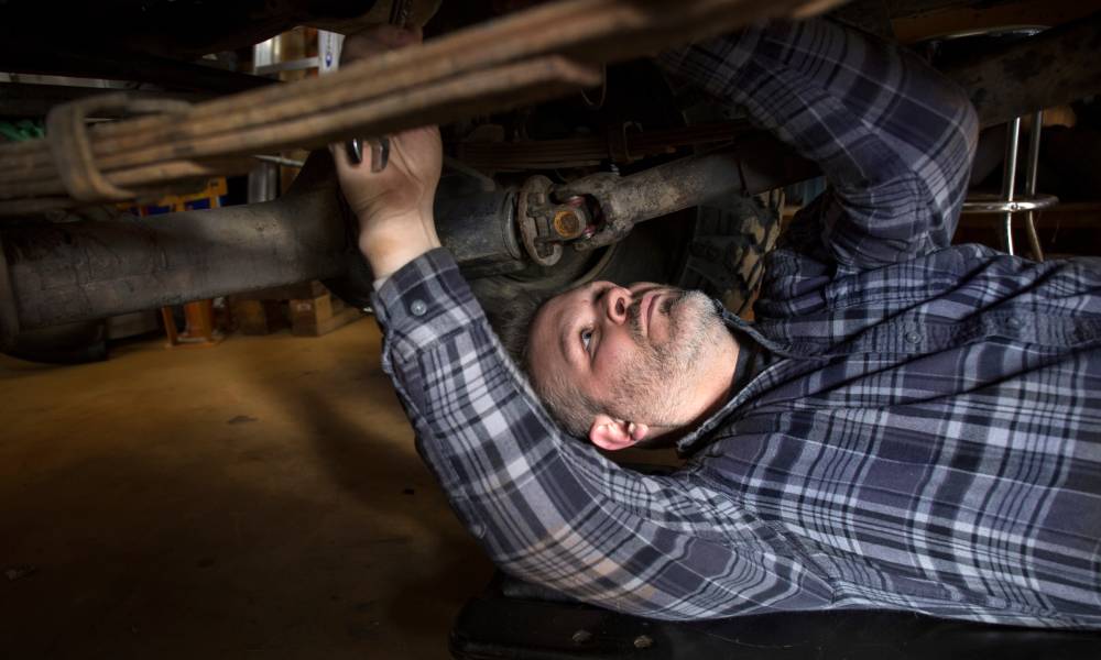 A man in a plaid button-down shirt lies on his back beneath the vehicle’s transmission to fix a transfer case issue.
