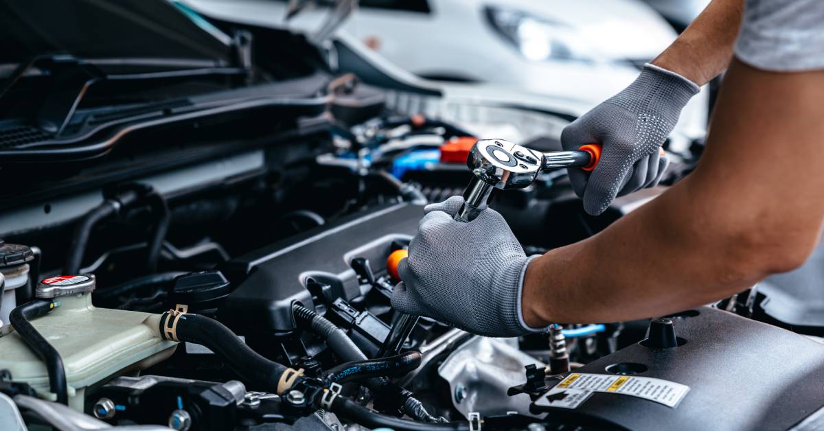 The hood of a car is open. A mechanic wears textured gloves and uses a wrench to tighten components on the transmission.