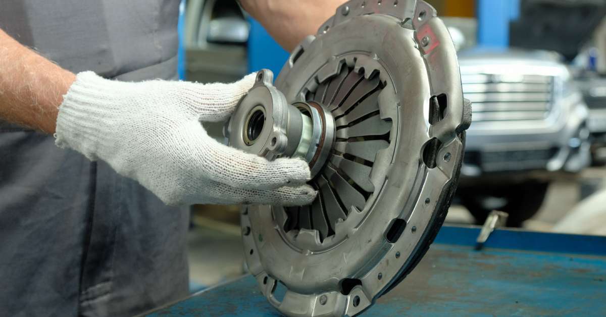 In an auto repair shop, a mechanic wearing gray overalls and white gloves examines a new clutch on a metal table.