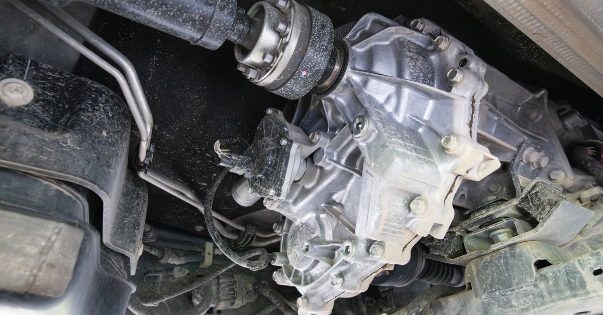 A transfer case sits underneath an SUV. The component has a silver metal housing that is slightly dirty.