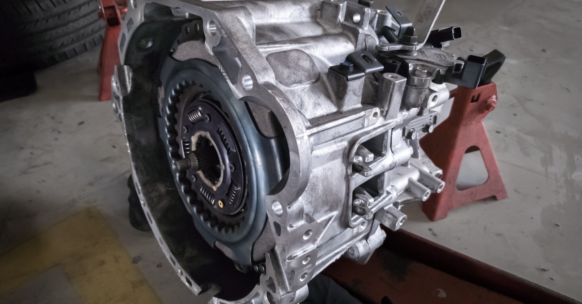 A close-up of a car transmission features a silver casing. It stands on adjustable legs on a garage floor.