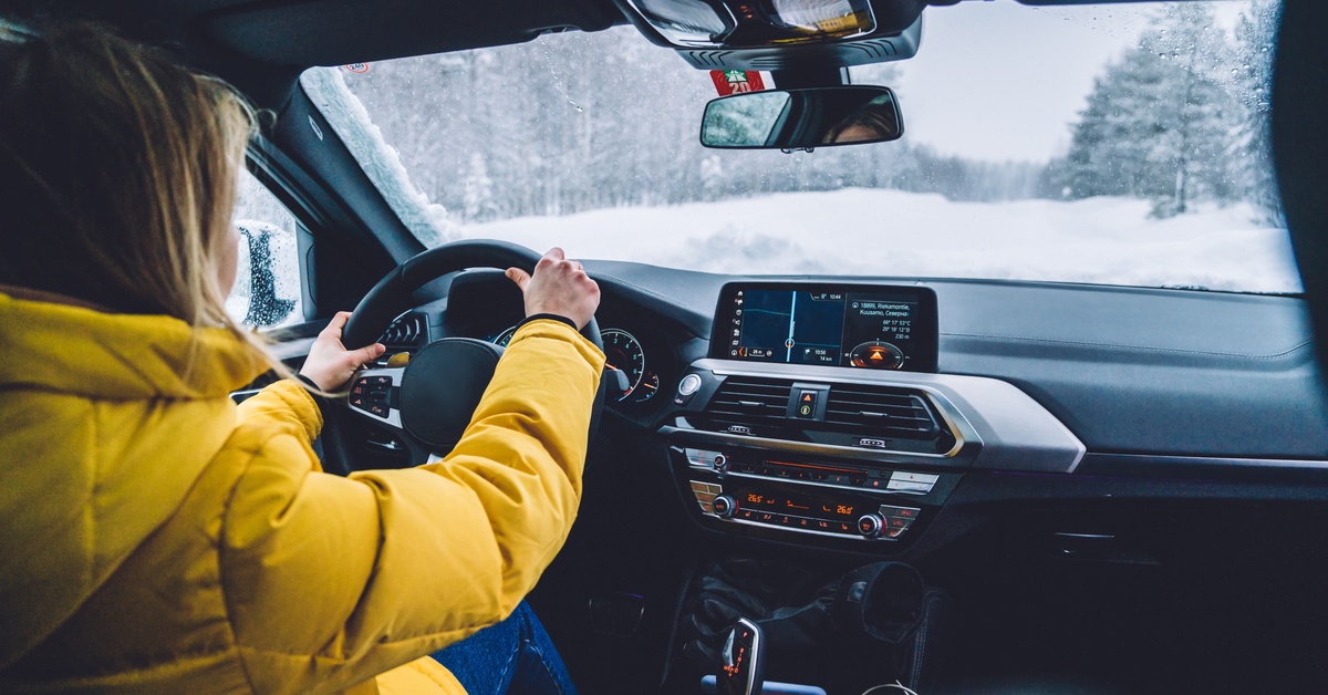 A woman wears a puffy yellow winter jacket as she drives her car. A snow-covered road is visible through the windshield.