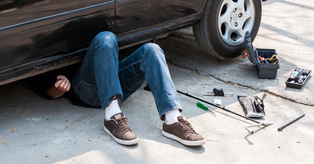 A person lies beneath a black car on a concrete driveway, with scattered tools and toolboxes nearby.