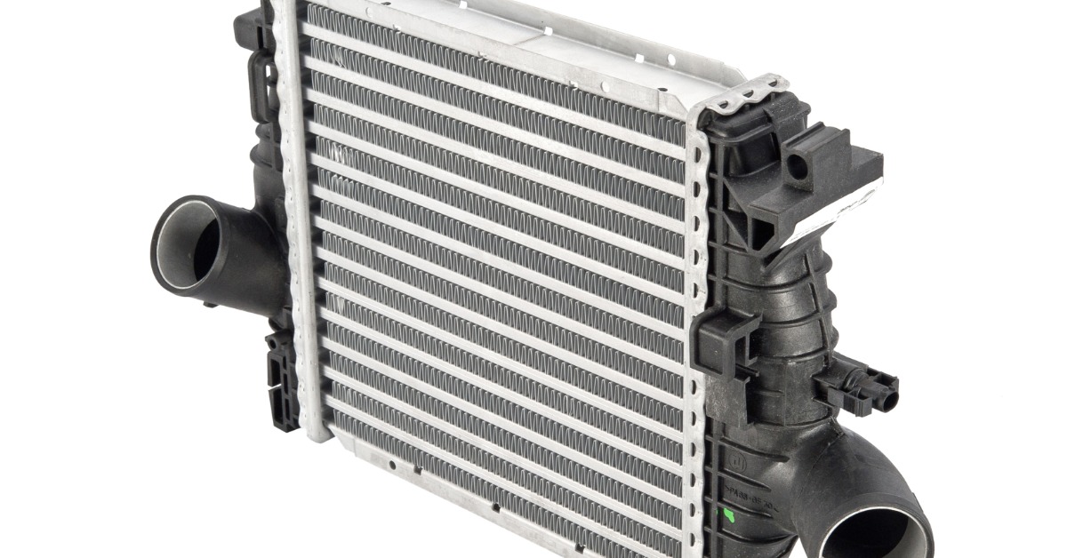 A car's silver radiator intercooler with metal cooling fins sits isolated on a white background. Tubes extend outward.